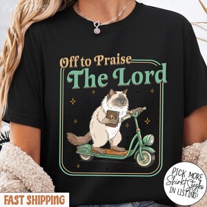 May include: Black t-shirt featuring a cartoon cat riding a green scooter and holding a book. The text "Off to Praise The Lord" is above the cat. The design includes small star accents and a decorative border.