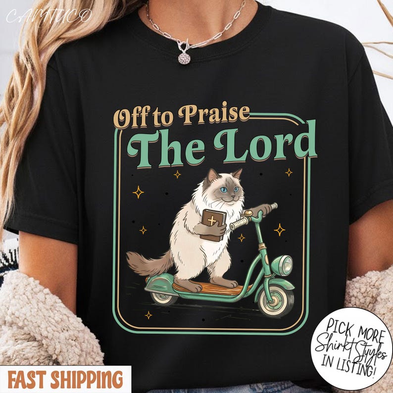 May include: Black t-shirt featuring a cartoon cat riding a green scooter and holding a book. The text "Off to Praise The Lord" is above the cat. The design includes small star accents and a decorative border.