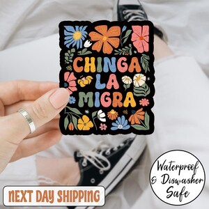 May include: A black sticker with colorful floral designs and the text "CHINGA LA MIGRA" in various colors. The sticker is surrounded by flowers in shades of orange, pink, blue, and yellow. The sticker is labeled "Waterproof & Dishwasher Safe".