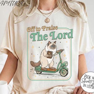 May include: Cream-colored t-shirt featuring a cartoon cat riding a green scooter and holding a book with a cross. The text "Off to Praise The Lord" is above the cat. The shirt has a round neck and short sleeves.