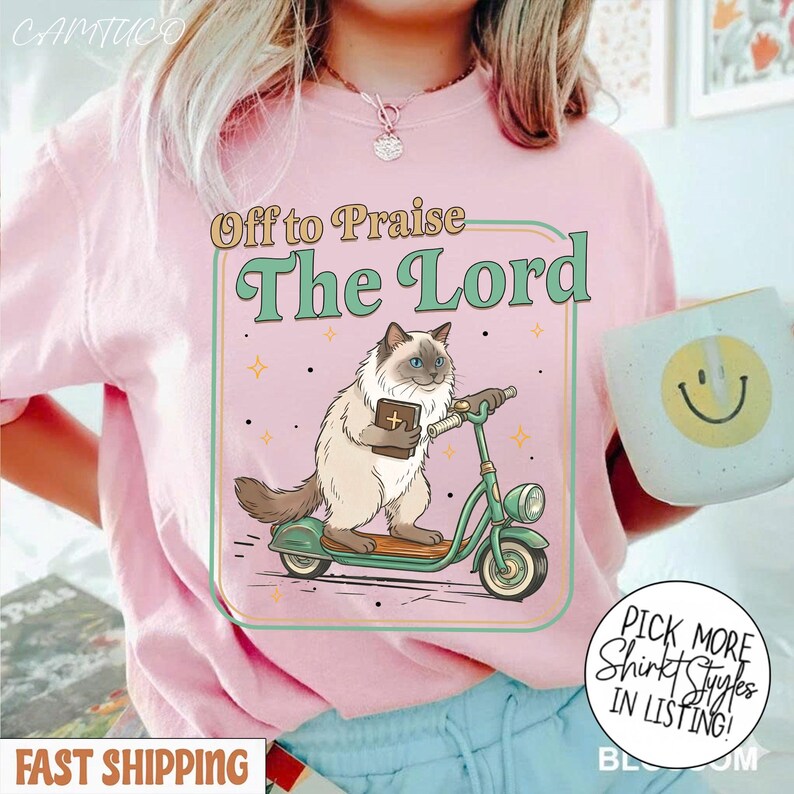 May include: Pink t-shirt with a graphic of a cat riding a scooter and holding a book, with the text "Off to Praise The Lord." The design is in a retro style. A mug with a smiley face is visible.