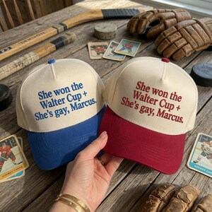 May include: Two baseball caps with cream-colored crowns and colored brims. One cap has a blue brim and blue embroidered text reading She won the Walter Cup + She's gay, Marcus. The other cap has a maroon brim and maroon text reading the same.