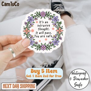 Pode incluir: Um autocolante branco com um design de grinalda floral em tons de roxo, rosa e verde. O autocolante tem o texto "It's an intrusive thought. It will pass. You are safe." O autocolante é segurado por uma mão.