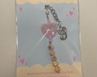 Puede incluir: Encanto de corazón rosa con brillo y un encanto de nota musical plateada unido a un llavero de cuentas rosas con el nombre "AYANA" escrito en letras doradas. El llavero está empaquetado en una tarjeta blanca con el texto "MOOMOO CREATIONS" y el sitio web "moo mooartscreations.etsy.com" y el nombre de usuario de las redes sociales "@moo moosproductsandco".
