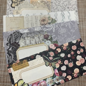 May include: A stack of five decorative envelopes with floral and bird patterns. The top envelope has a white background with black floral designs. The second envelope has a light blue background with white floral designs. The third envelope has a gray background with silver floral designs. The fourth envelope has a black background with pink and white floral designs. The bottom envelope has a white background with a gold border and a floral design. The text "TOUT-PARIS" is visible on the second envelope.