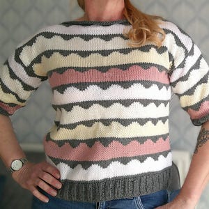 Puede incluir: Un suéter tejido con un patrón de rayas en blanco, gris, rosa y amarillo. El suéter tiene mangas cortas y cuello barco. Los puños y el dobladillo son grises.