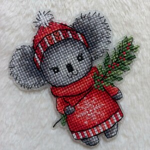 May include: A gray koala wearing a red and white striped hat and a red sweater with a snowflake design. The koala is holding a green sprig of holly.