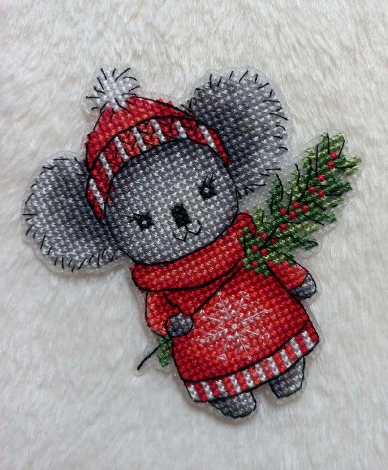May include: A gray koala wearing a red and white striped hat and a red sweater with a snowflake design. The koala is holding a green sprig of holly.