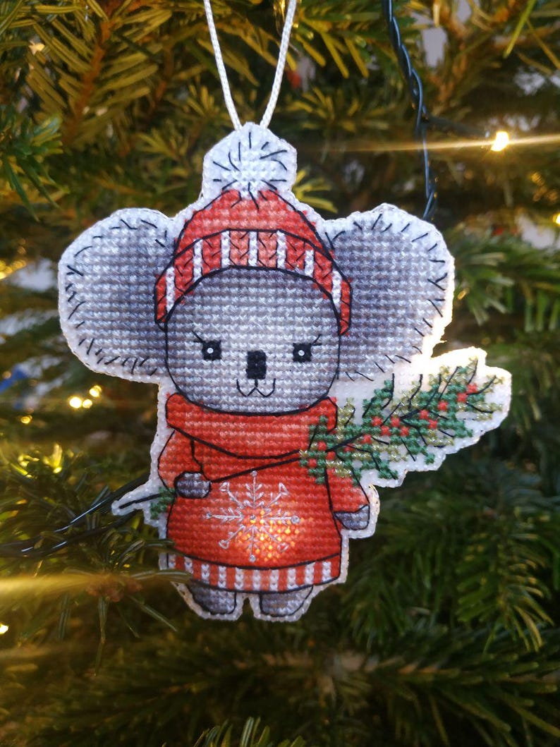May include: A gray koala wearing a red and white striped hat and a red sweater with a snowflake design. The koala is holding a green sprig of holly in its right paw. The ornament is hanging on a Christmas tree.