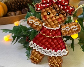 May include: A handmade gingerbread woman ornament with a red dress and a red bow with white polka dots. The ornament is made with cross-stitch embroidery and has a gold beaded necklace. The background includes holly and fairy lights.
