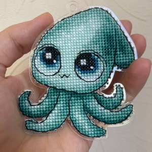 May include: A teal blue crocheted squid with large eyes and a cute expression. The squid is attached to a clear plastic keychain.