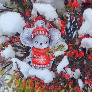 May include: A gray and white cross-stitch ornament of a mouse wearing a red and white striped hat and a red sweater with a snowflake design. The ornament is holding a green sprig of holly. The ornament is surrounded by snow-covered branches and red berries.