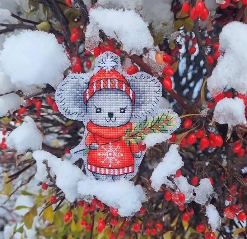May include: A gray and white cross-stitch ornament of a mouse wearing a red and white striped hat and a red sweater with a snowflake design. The ornament is holding a green sprig of holly. The ornament is surrounded by snow-covered branches and red berries.