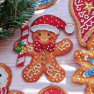 May include: A gingerbread man Christmas ornament with a red and white hat, holding a candy cane. The ornament is made of cross-stitch fabric and is decorated with white beads.