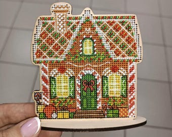 May include: A wooden gingerbread house ornament with a white icing roof, green windows, and a red door. The house is decorated with red and green candy and has a white base.