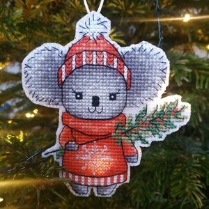 May include: A gray koala wearing a red and white striped hat and a red sweater with a snowflake design. The koala is holding a green sprig of holly in its right paw.