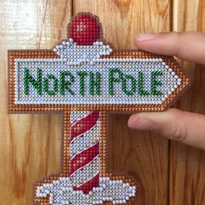 May include: A cross-stitch signpost pointing to the North Pole. The sign is brown with white beaded edges and green text. The pole is red and white striped. A red ball sits atop the sign.