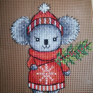 May include: A gray and white koala wearing a red and white striped hat and a red sweater with a white snowflake design. The koala is holding a green sprig of holly with red berries.