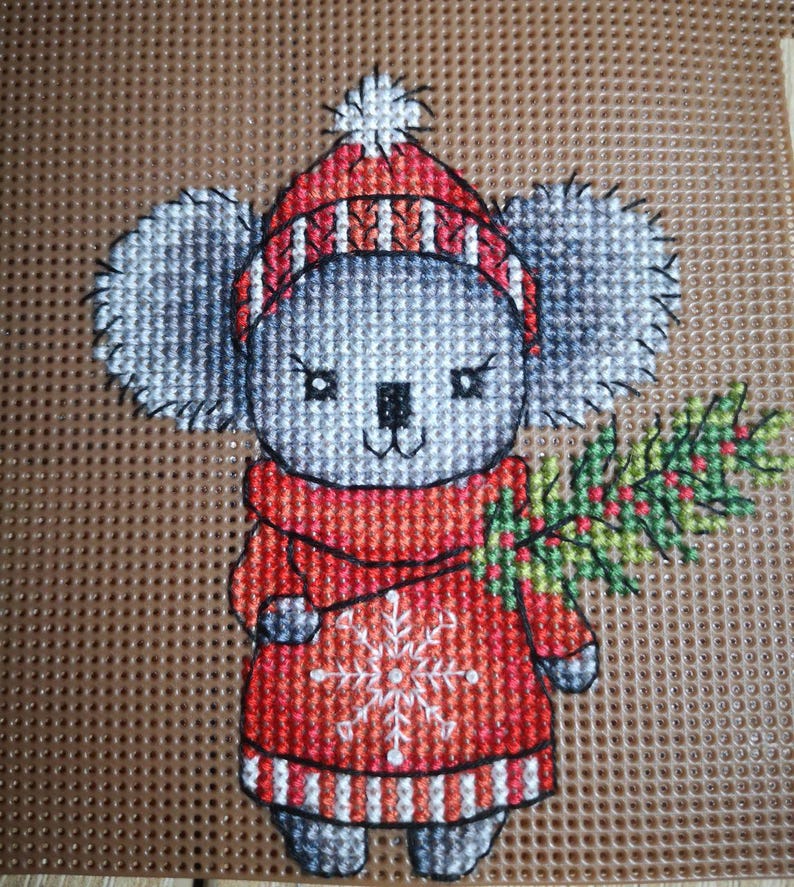 May include: A gray and white koala wearing a red and white striped hat and a red sweater with a white snowflake design. The koala is holding a green sprig of holly with red berries.