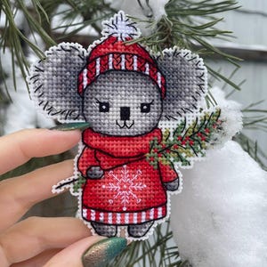 May include: A cross-stitch ornament of a koala wearing a red and white striped hat and scarf, holding a sprig of holly. The koala is wearing a red sweater with a white snowflake design.