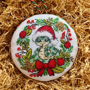 May include: A round, embroidered Christmas ornament featuring a green snake wearing a Santa hat. The snake is surrounded by a wreath of red berries, green leaves, and a red bow. The ornament has a green and red hanging cord.