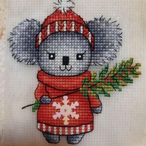 May include: A cross-stitch pattern of a koala wearing a red and white striped hat and a red scarf with a snowflake on it. The koala is holding a green branch with red berries.