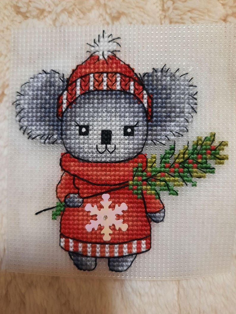 May include: A cross-stitch pattern of a koala wearing a red and white striped hat and a red scarf with a snowflake on it. The koala is holding a green branch with red berries.