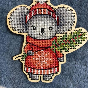 May include: A gray koala wearing a red and white striped hat and sweater with a snowflake design. The koala is holding a green sprig of holly with red berries. The ornament is made of wood and is perfect for Christmas decor.