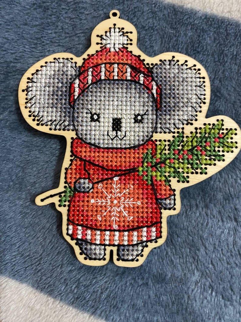 May include: A gray koala wearing a red and white striped hat and sweater with a snowflake design. The koala is holding a green sprig of holly with red berries. The ornament is made of wood and is perfect for Christmas decor.