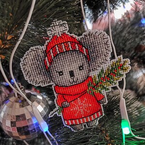 May include: A gray and white cross-stitch ornament of a koala wearing a red and white striped hat and sweater, holding a green sprig of holly. The ornament is hanging on a Christmas tree.