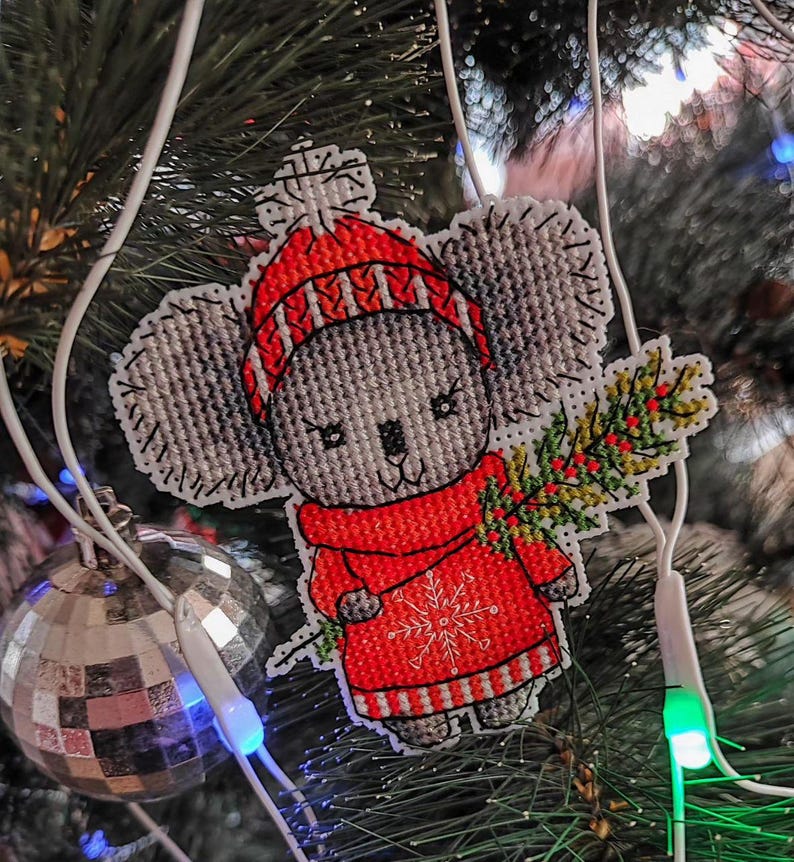 May include: A gray and white cross-stitch ornament of a koala wearing a red and white striped hat and sweater, holding a green sprig of holly. The ornament is hanging on a Christmas tree.