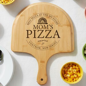 May include: A wooden pizza peel with the text "MOM'S PIZZA" and "VENTNOR, NEW JERSEY" is surrounded by small bowls of pizza toppings and a set of silverware on a white plate. The peel has a handle and a rounded top.