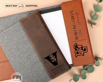Puede incluir: Una carpeta de tela gris con una solapa de cuero marrón oscuro con el logotipo del Vanderbilt University Medical Center y una solapa de cuero marrón claro con el logotipo de la Bowling Green State University, ambas con blocs de papel rayado.