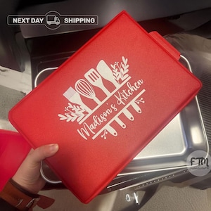 May include: A rectangular red lid for a baking pan featuring a white graphic design of kitchen utensils including a whisk, spatula, and brush, with the text Madison's Kitchen printed in a script font across the center of the design.