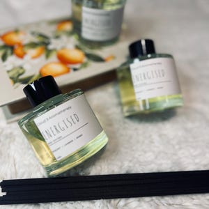 May include: Three glass aromatherapy bottles with black caps and the word "ENERGISED" on the label. The bottles are filled with a light green liquid. A stack of black diffuser sticks is in the foreground. A decorative box with orange fruit is in the background.