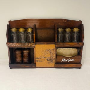 May include: A vintage wooden kitchen organizer with multiple compartments. It features glass spice jars with gold lids, a recipe box labeled "Recipes", and a corkboard with a landscape design. Two wooden salt and pepper grinders are also visible.