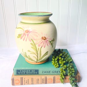 May include: A cream-colored ceramic vase with a crackle glaze finish, featuring hand-painted pink daisy-like flowers and green foliage. The vase has a wide, rounded body, a flared rim with a green interior, and a brown base.