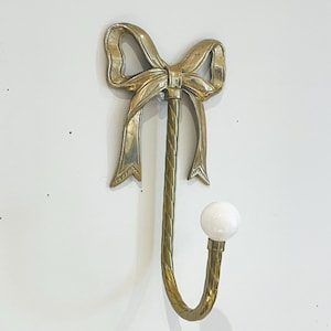 Könnte beinhalten: Ein dekorativer Wandhaken in Form einer Schleife, aus messingfarbenem Metall. Der Haken hat ein gebogenes Design mit einem weißen, kugelförmigen Knauf am Ende. Die Schleife ist mit strukturierten Kanten versehen.