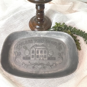 May include: A rectangular, pewter-toned tray with rounded corners. The tray is embossed with the phrase "BLESS THIS HOUS" and a detailed image of a house, trees, and floral accents. A wooden candlestick is in the background.