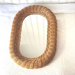 May include: An oval-shaped wall mirror featuring a thick, woven light brown wicker frame. The mirror has a smooth, reflective glass surface surrounded by a textured, braided natural reed border that creates a continuous, rounded edge design.
