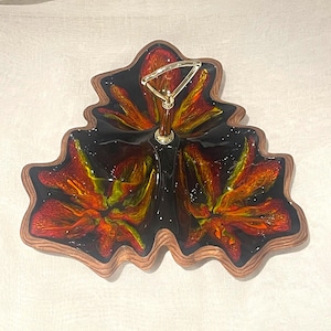 Op de afbeelding: Decoratieve serveerschaal in de vorm van een blad. De glanzende binnenkant heeft levendige rode, oranje en gele bloemmotieven. Met houten rand en goudkleurige handgreep. Uniek voor het serveren van snacks of het presenteren van items, ongeveer 30 cm breed.