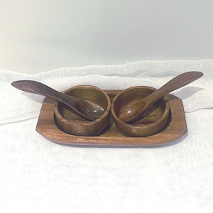 May include: A wooden serving set featuring two small bowls and two spoons, all resting on a rectangular tray. The set is made of a warm-toned wood, perfect for serving dips or condiments. The spoons are curved to fit the bowls.