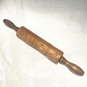 May include: A wooden rolling pin with carved floral and geometric designs. The rolling pin has dark brown handles and a cylindrical rolling surface. The surface is decorated with carved patterns, including flowers and stars.