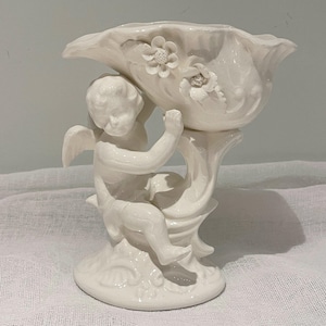 Puede incluir: Una figura de querubín de cerámica blanca sosteniendo un cuenco decorativo. El cuenco tiene detalles florales y un borde festoneado. El querubín tiene alas y está sentado sobre una base de pedestal. Una pieza decorativa para el hogar.