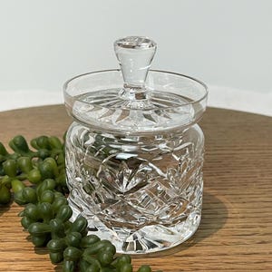 Könnte beinhalten: Ein klarer, geschliffener Glasbehälter mit Deckel und dekorativem Knauf. Der Behälter weist ein Diamantenmuster auf und steht auf einer Holzoberfläche. Der Behälter wird wahrscheinlich zur Aufbewahrung kleiner Gegenstände oder als Dekorationsstück verwendet.