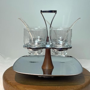 Könnte beinhalten: Ein silberfarbenes Servierset mit zwei transparenten Glasschalen, jeweils mit einem kleinen Löffel. Die Schalen stehen auf einem silbernen Tablett mit Holzsockel und Griff. Das Set ist für das Servieren von Gewürzen oder kleinen Portionen konzipiert.