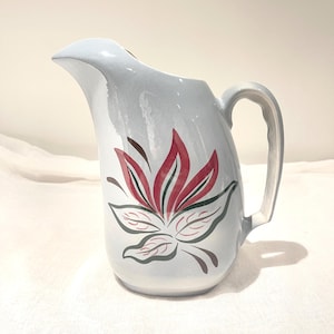 Puede incluir: Jarra de cerámica azul claro con asa y pico vertedor. Presenta un gran diseño floral en rojo, verde y marrón. Esta jarra se utiliza probablemente para servir bebidas.