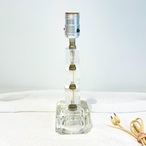 May include: A clear glass table lamp with a square base and a stacked cube design. The lamp has a silver-colored bulb socket and a tan electrical cord. The lamp measures approximately 12 inches tall.