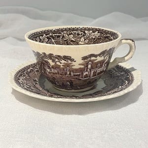 May include: A vintage teacup and saucer set. The cup and saucer are cream-colored with a brown transferware design depicting a landscape scene. The cup has a handle and the saucer has a scalloped edge. The set is likely made of porcelain.