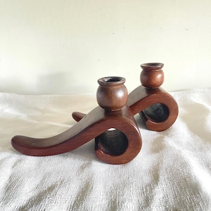 May include: Two wooden candle holders with a rich, dark brown finish. Each holder features a small, round cup for a candle and a curved, scroll-like design. The holders are placed on a white, textured surface.